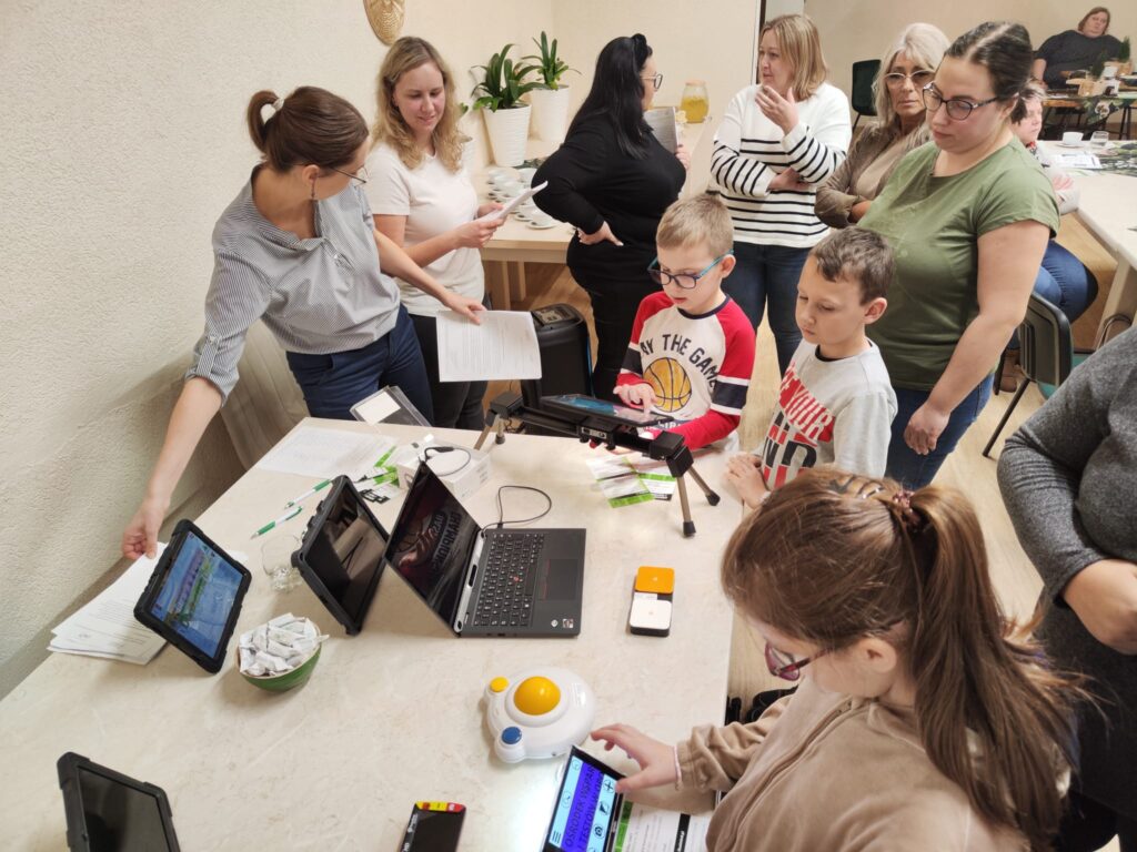 Grupa kobiet i dzieci stoi przy stole. Na stole są technologie asystujące: tablety z aplikacją Mówik, lupa elektroniczna, powiększone myszki komputerowe, powiększalnik elektroniczny Spider