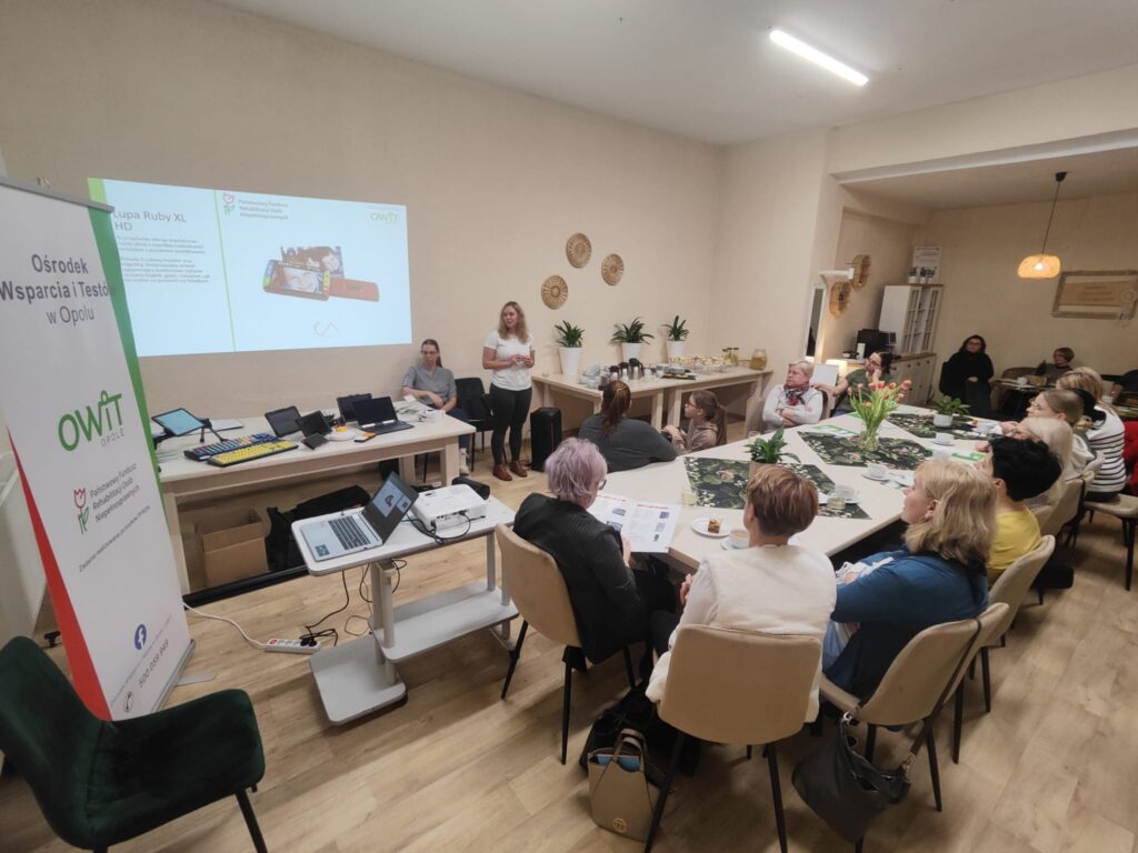 Sala z długim stołem. Przy stole siedzą kobiety i dziewczynka. Na ścianie wyświetlona prezentacja o lupie elektronicznej Ruby XL. Obok ściany dwie pracownice OWiT w Opolu