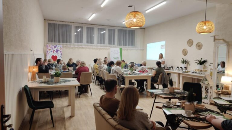 Duża sala ze stołami. Przy stołach siedzą mężczyźni, kobiety i dzieci. Pracownica OWiT stoi przy ścianie. Na ścianie za nią wyświetlona prezentacja. W tle rollap z napisem OWiT