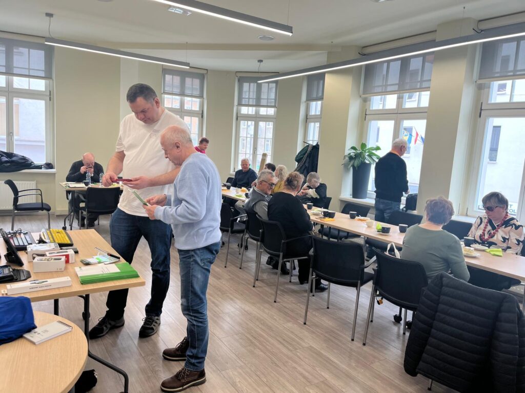 W sali przy stole z technologiami asystującymi stoi dwóch mężczyzn. Pracownik OWiT prezentuje mężczyźnie lupę elektroniczną. W tle przy stołąch siedzą członkowie CROSS i prowadzą rozmowy