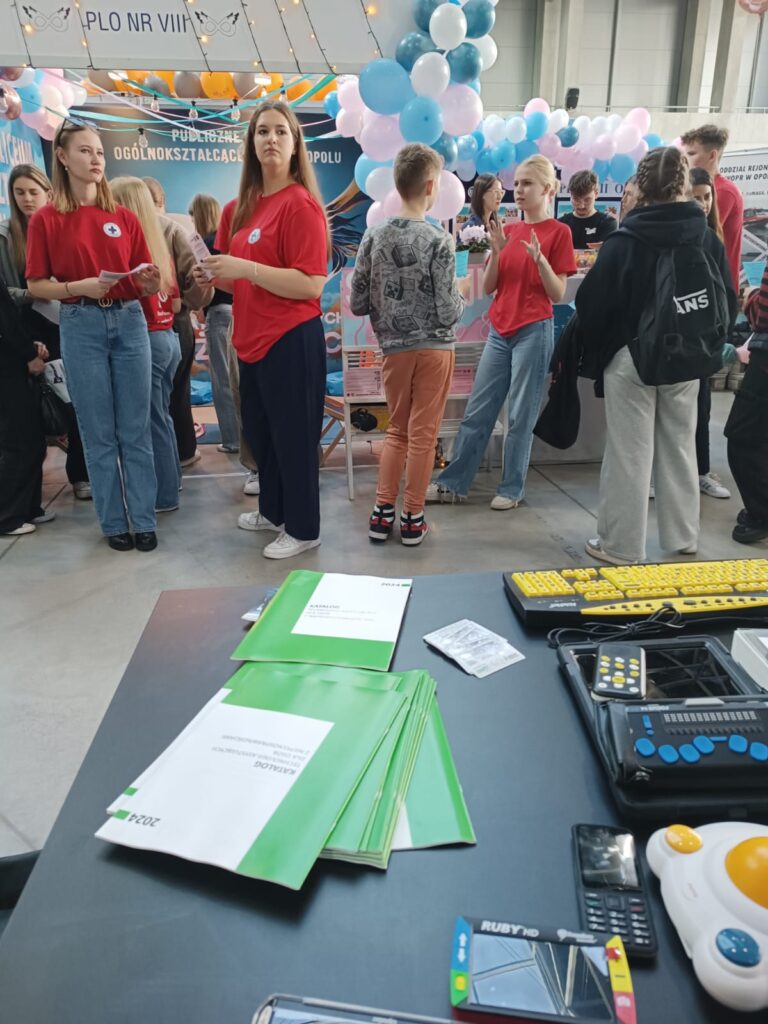 Stoisko OWiT Opole. Na stole rozłożone technologie asystujące, katalogi sprzętu dostępnego w OWiT. W głębi kilkoro uczennic i uczniów zwiedzających Targi.
