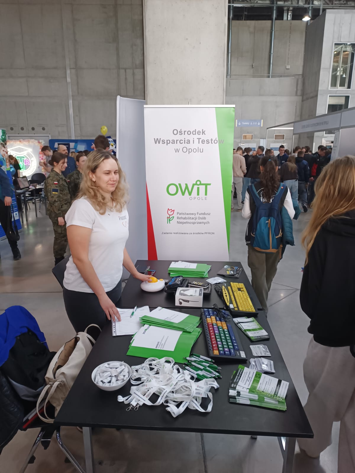 Stoisko OWiT Opole. Na stołach rozłożone technologie asystujące, katalogi sprzętu dostępnego w OWiT, ulotki, smycze reklamowe, długopisy i krówki. W głębi rollup reklamowy OWiT z napisem Ośrodek Wsparcia i Testów w Opolu oraz logo OWiT i PFRON.
