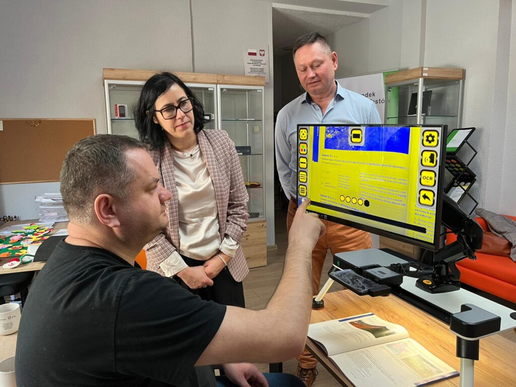 Pracownik OWiT siedzi przy stole przed powiększalnikiem Pulchra Es. Zaznacza pola na ekranie. Obok niego stoją kobieta i mężczyzna, przyglądają się ekranowi