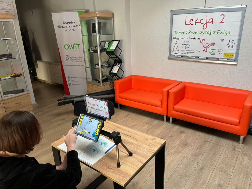 Pracownica OWiT siedzi przy stole przed powiększalnikiem Exigo. Zaznacza na ekranie dostępne funkcje
