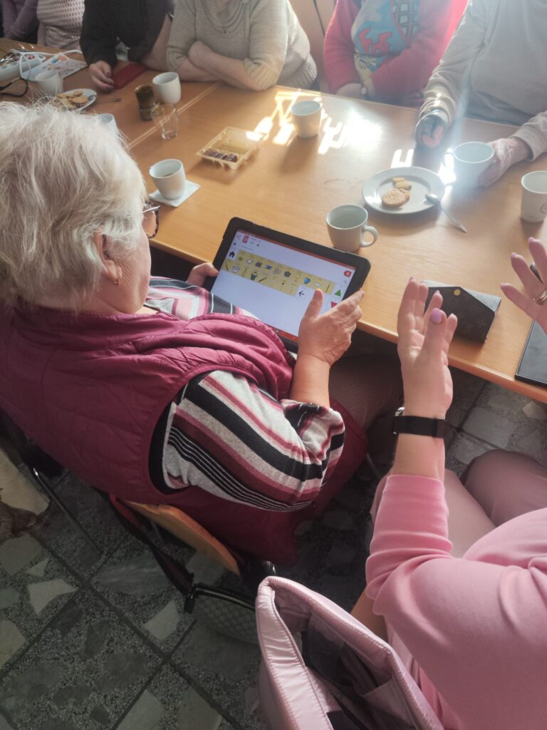 Przy stole siedzi kilka osób. Kobieta trzyma w rękach tablet z oprogramowaniem Mówik