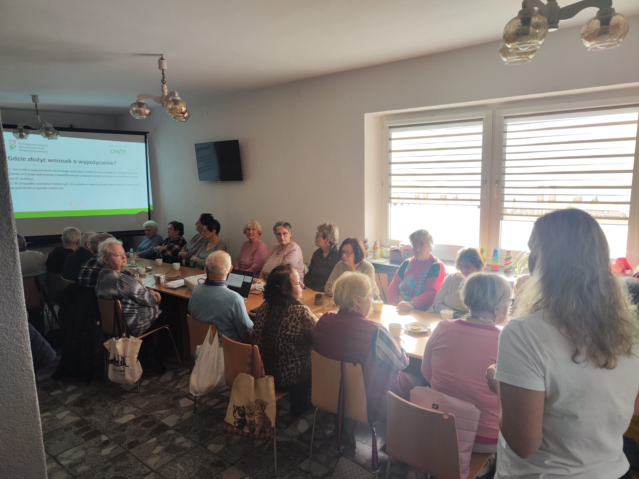 Sala z długim stole. Przy stole siedzi około dwadziestki kobiet i mężczyzni. Za nimi na ścianie wyświetlona prezentacja z napisem Gdzie złożyć wniosek o wypożyczenie.