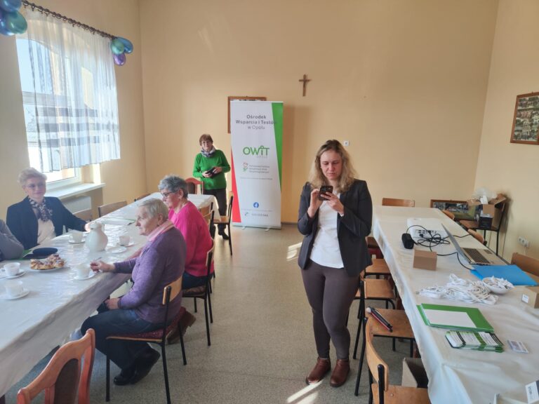Dwa długie stoły. Przy stole po lewej stronie siedzi kilka osób, na stole po prawej stronie rozłożone katalogi i ulotki OWiT. Na pierwszym planie pracownica OWiT prezentuje smartfon