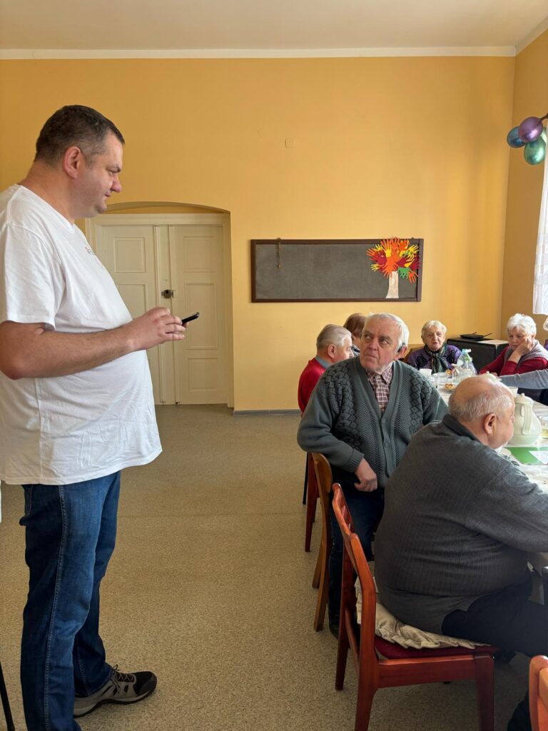 Po prawej stronie stół, przy którym siedzi kilka osób. Po lewej stronie stoi pracownik OWiT i rozmawia z mężczyzną siedzącym przy stole
