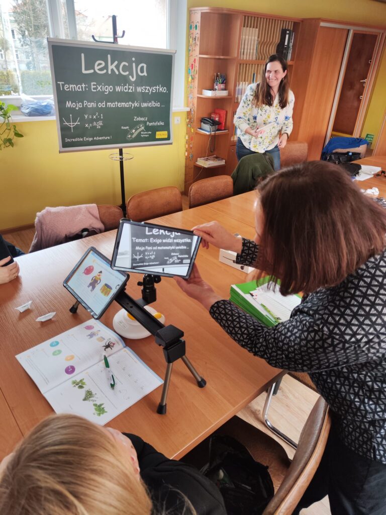 Na stole leży rozłożony podręcznik. Nad nim stoi powiększalnik Exigo. Pracownica OWiT pokazuje ustawienia powiększalnika. Na jednym tablecie widać powiększoną stronę podręcznika. Na drugim tablecie widać powiększoną tablicę wiszącą na stojaku w odległości około 3 metrów od powiększalnika
