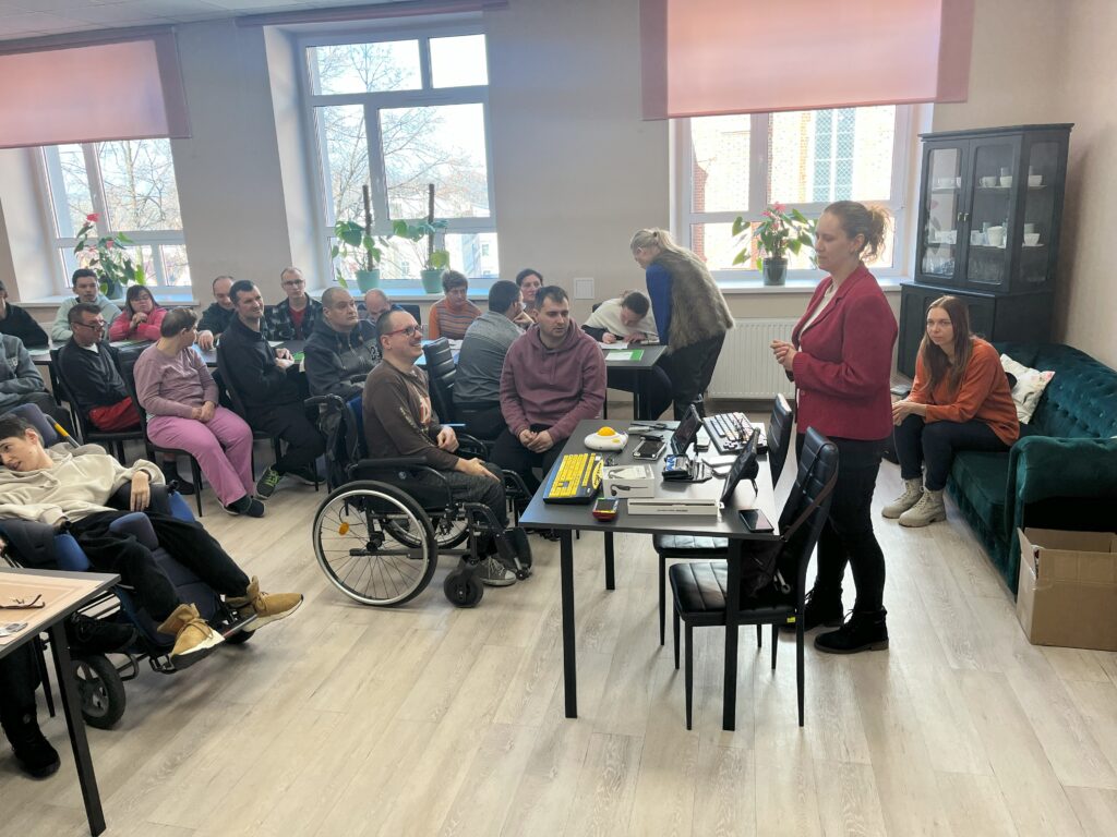 Na sali widocznych około 20 uczestników Środowiskowego Domu Samopomocy. Niektórzy siedzą na krzesłach, dwie osoby siedzą na wózkach. Na środku stoi pracownica OWiT i opowiada o technologiach asystujących, które leżą przed nią na stole