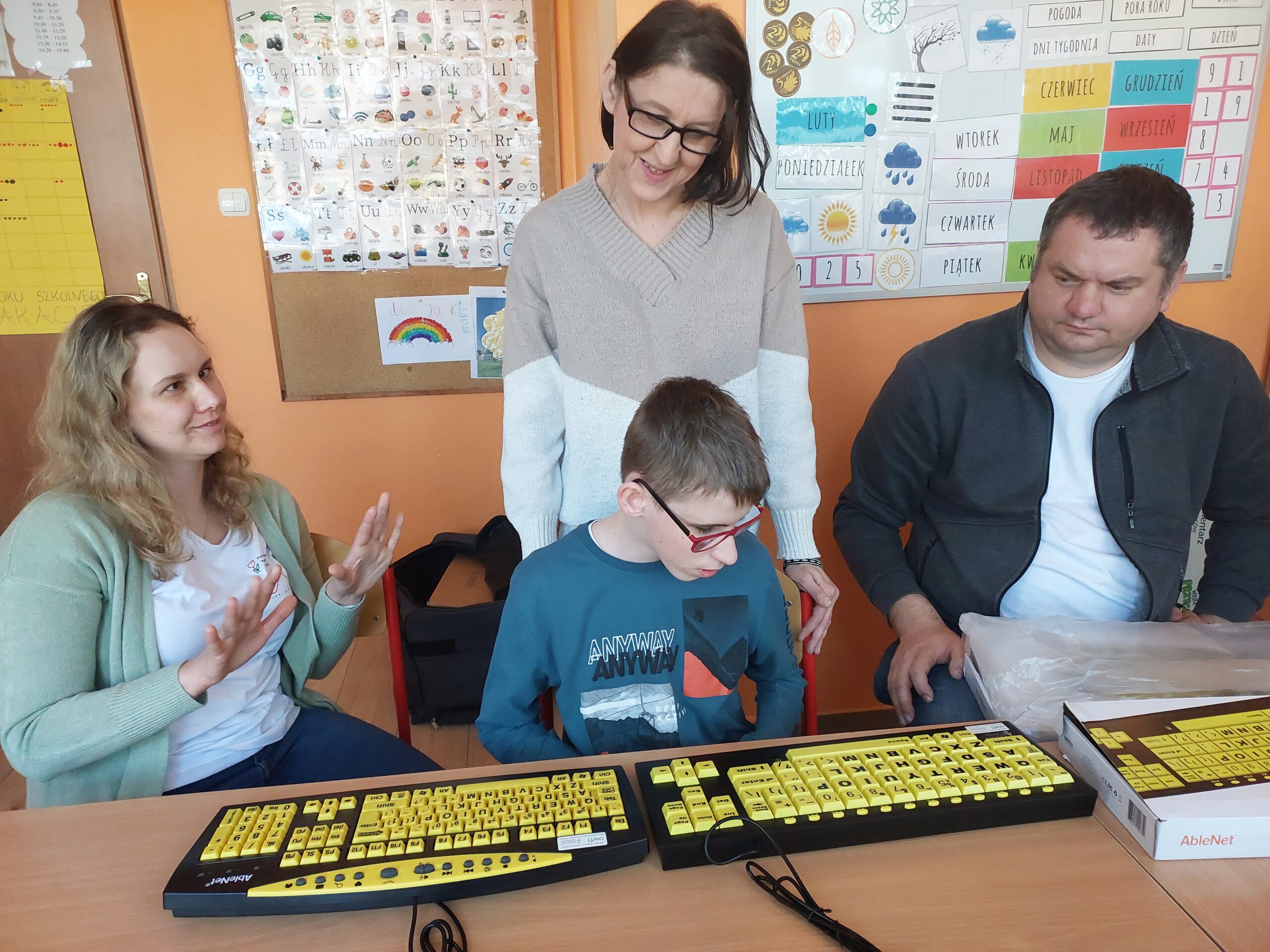Trzy osoby siedzą przy stole, kobieta stoi za nimi. Przy stole w środku chłopiec w okularach patrzy na specjalistyczną klawiaturę komputerową z powiększonymi klawiszami. Klawisze są w żółtym kolorze, na nich litery, cyfry i napisy w czarnym kolorze. Po obu stronach chłopca siedzą pracownicy OWiT, pracownica po lewej stronie gestykuluje.