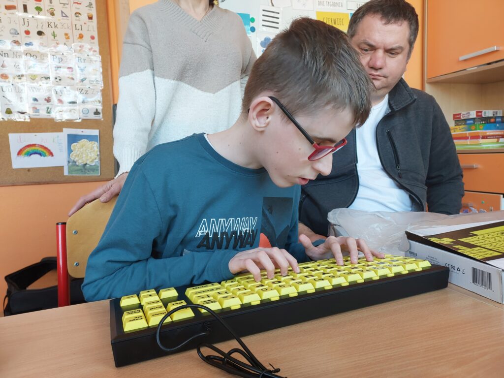 Chłopiec w okularach siedzi przy stole, palcami dotyka klawiszy specjalistycznej klawiatury. Klawisze są w żółtym kolorze, na nich litery, cyfry i napisy w czarnym kolorze.