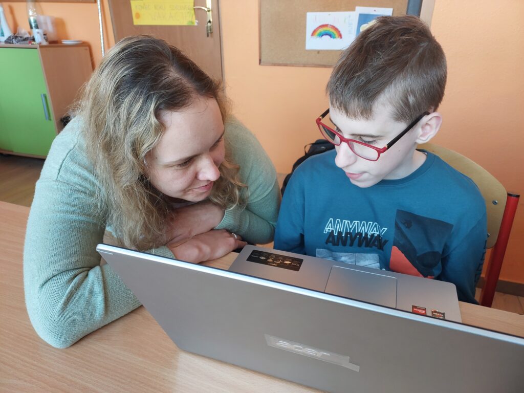 Przy stole po prawej stronie siedzi chłopiec w okularach. Obok niego po lewej stronie siedzi pracownica OWiT. Oboje patrzą na otwarty laptop, który stoi przed nimi.