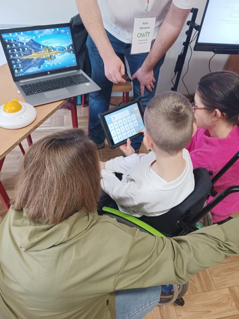 Tyłem do nas trzy osoby. W środku na wózku siedzi kilkuletni chłopiec, po jego obu stronach kucają dwie kobiety. Chłopiec patrzy w ekran tabletu z oprogramowaniem Mówik