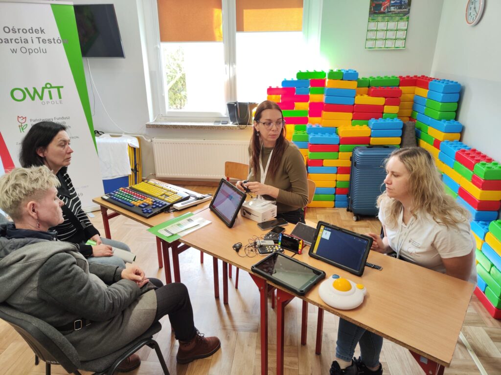 Po obu stronach stołu siedzą 4 kobiety. Dwie pracownice OWiT prezentują technologie asystujące dwóm paniom. Na stole rozłożone  tablety z oprogramowaniem Mówik i Grid,, duża mysz komputerowa, dwie specjalistyczne klawiatury, lupy powiększające