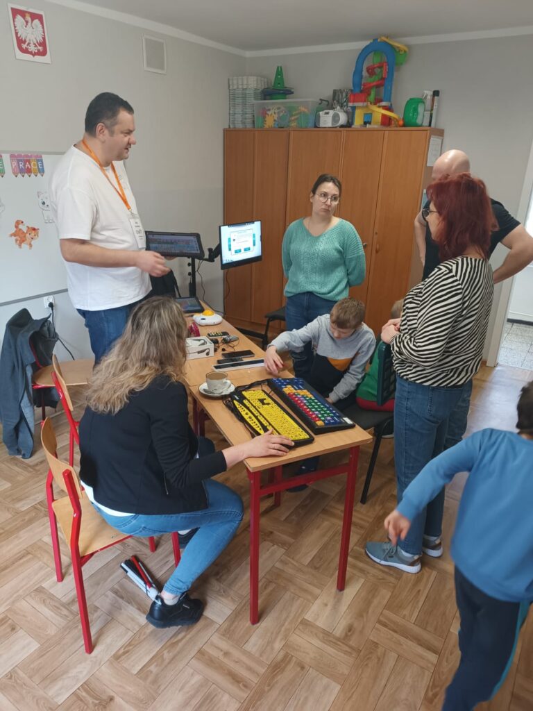 Grupa 7 osób, pracownica OWiT i dwoje dzieci siedzą przy stole. Pozostałe osoby stoją. Pracownik OWiT rozmawia z ze stojącymi osobami. W tle stoi Cyberoko