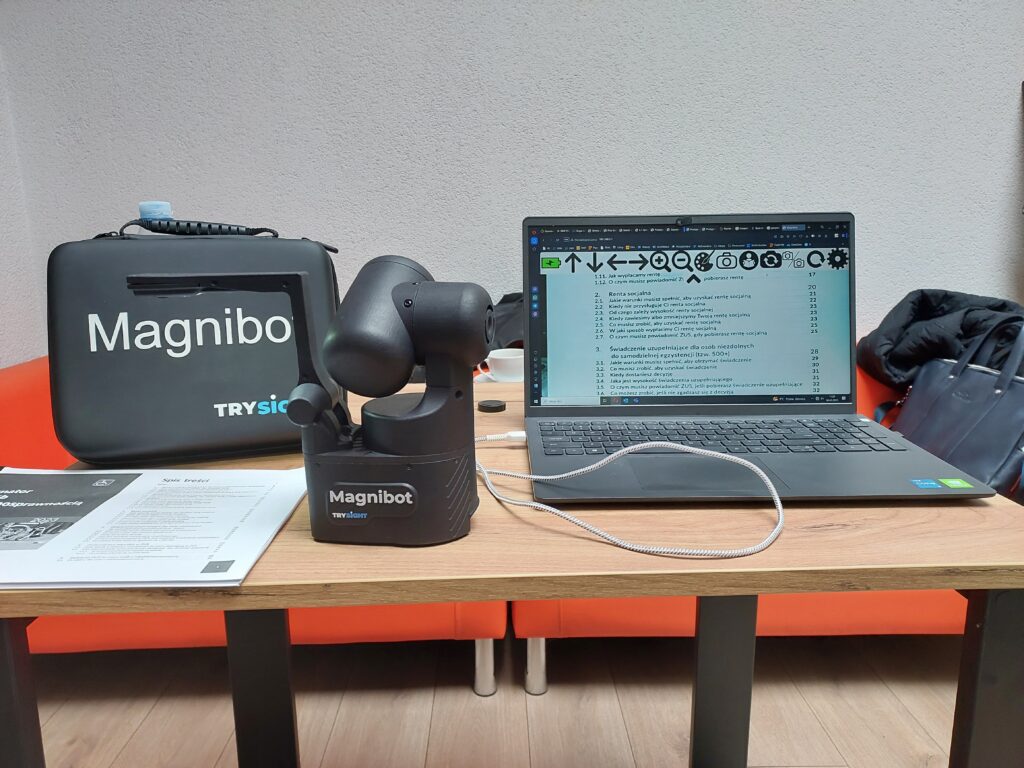 Magnibot: na biurku stoi Magnibot, po jego lewej stronie leży publikacja otwarta na spisie treści. Po prawej stronie Magnibota stoi otwarty laptop. Na ekranie laptopa widoczna powiększona spis treści publikacji.
