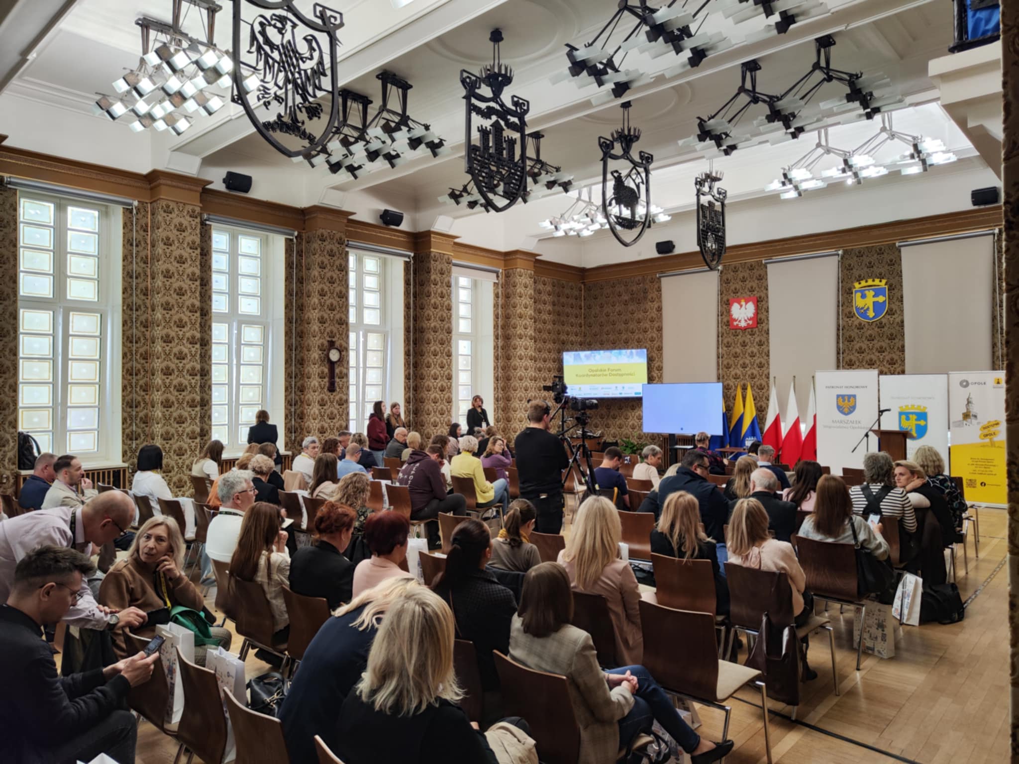 Duża sala wypełniona rzędami krzeseł. Na krzesłach siedzą kobiety i mężczyźni w różnym wieku. Z przodu Sali flagi polskie i opolskie, bannery z logo województwa opolskiego i miasta Opola, ekran z wyświetloną nazwą Opolskie Forum Koordynatorów Dostępności