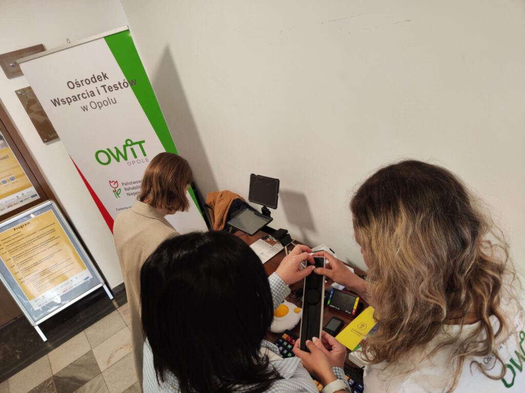 Po lewej stronie stoi potykacz z programem Forum. Obok stoi banner OWiT w Opolu. Po prawej stronie na stole stoją technologie asystujące: Exigo, switch, klawiatura specjalistyczna. Stół częściowo zasłonięty jest przez 3 osoby oglądające technologie