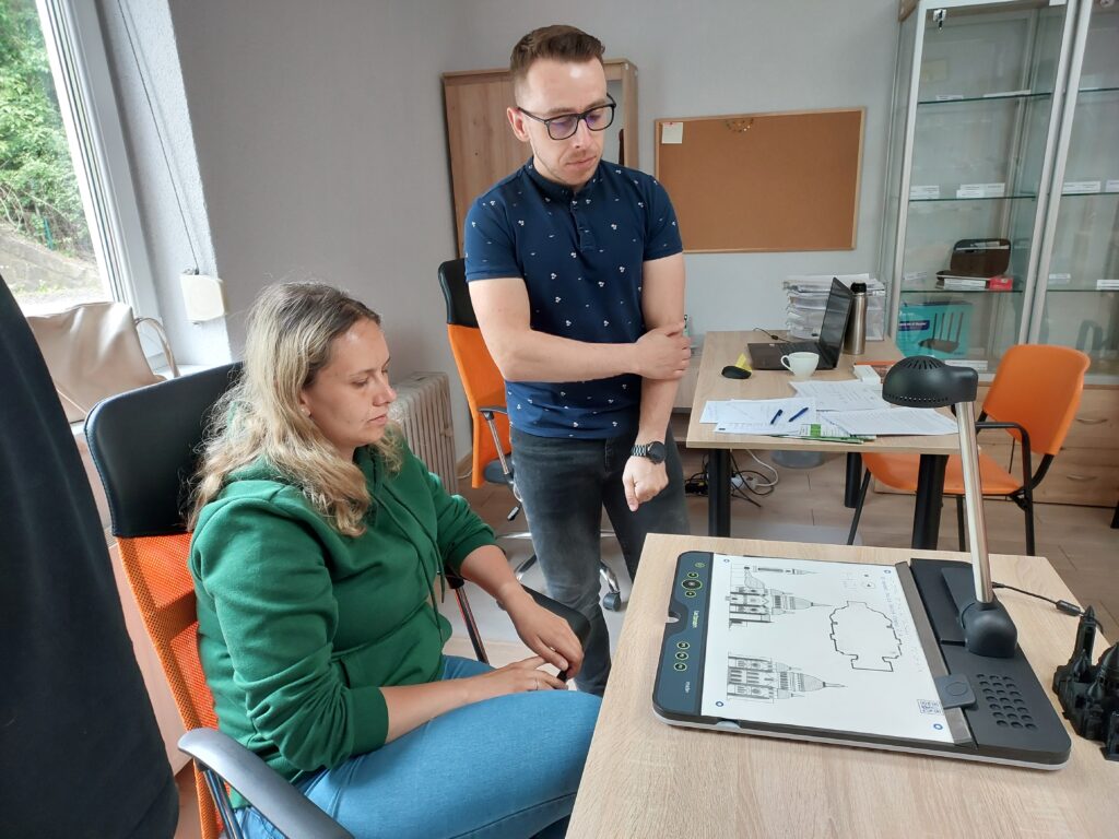 estujemy Tactonom: dwie osoby, kobieta siedzi przy biurku, przed nią na biurku stoi Tactonom. Za kobietą po prawej stronie stoi mężczyzna, patrzy na Tactonom stojący na biurku.