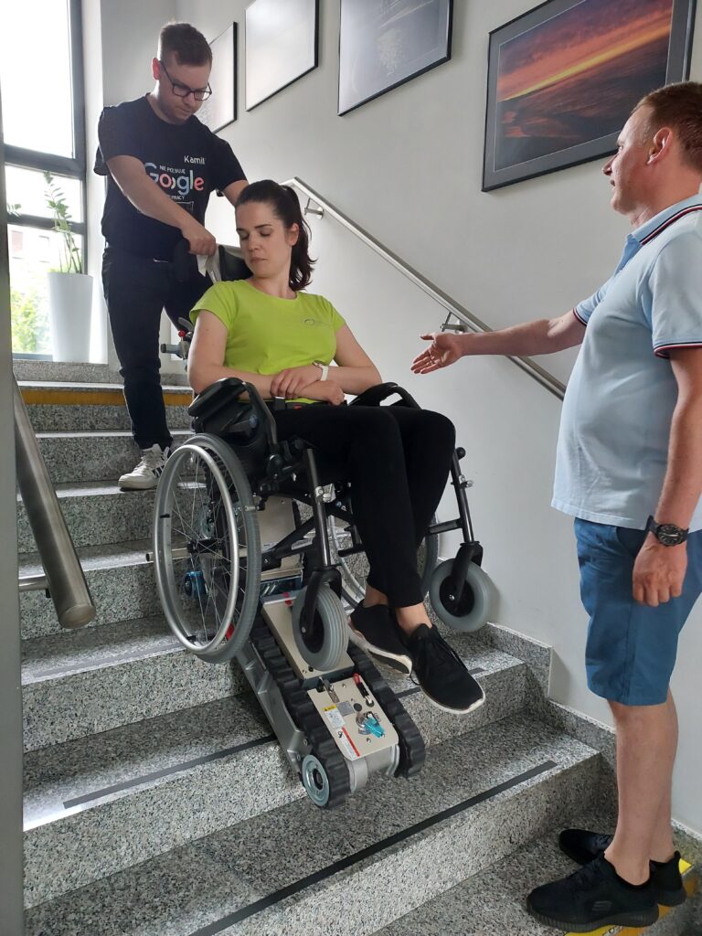 W połowie schodów znajduje się schodołaz którym kieruje młody mężczyzna. Na wózku na schodołazie siedzi młoda kobieta. Przed schodołazem stoi instruktor z prawą ręką wyciągniętą przed przed siebie.