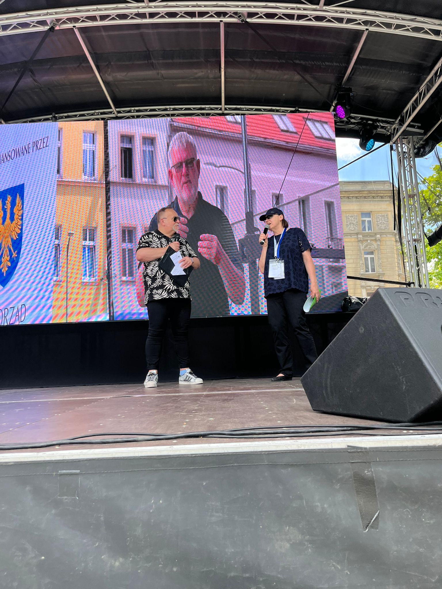 Na estradzie stoją dwie osoby. Po lewej mężczyzna z mikrofonem w ręku, po prawej przedstawicielka OWiT z mikrofonem w ręku. Na ekranie w tle tłumacz polskiego języka migowego.