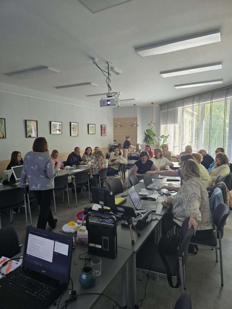 Grupa kobiet i mężczyzn widocznych siedzących przy stołach ustawionych w podkowę. Na pierwszym planie widoczna od tyłu prowadząca spotkanie.