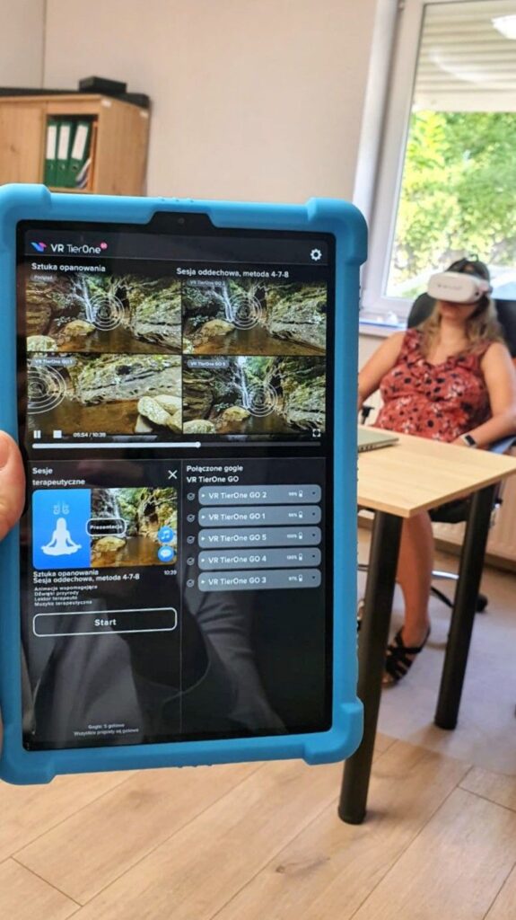 Na pierwszym planie tablet z zakładkami do kierowania goglami VR Tier One. W tle na fotelu biurowym siedzi kobieta z goglami założonymi na głowę.