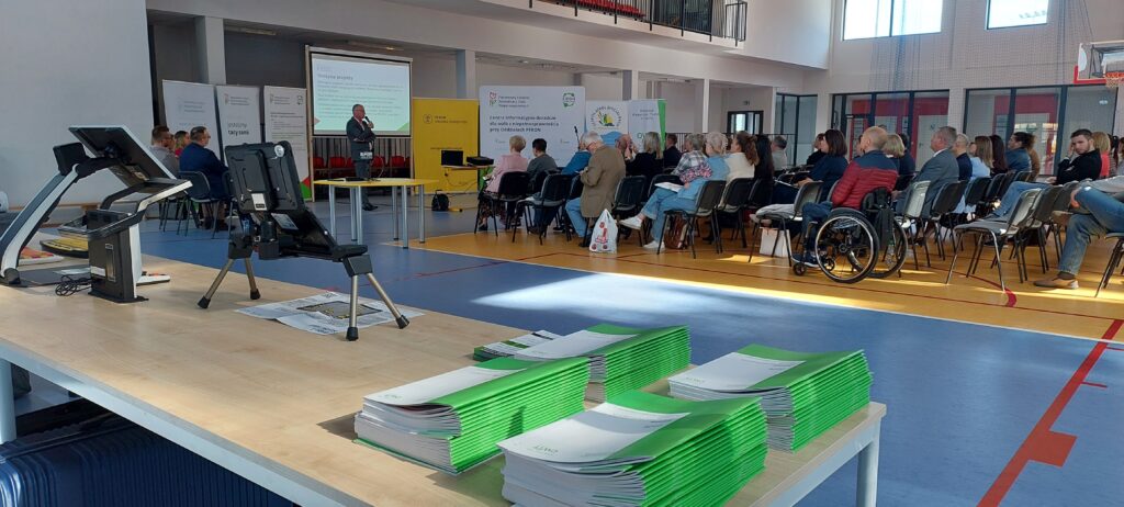 Duża sala. Na pierwszym planie stół z katalogami technologii asystujących na 2026 rok. W głbi rzędy krzeseł z siedzącymi na nich osobami. Jedna osoba siedzi na wózku. Po lewej stronie ekran, rollupy. Przed nimi stoi dyrektor opolskiego oddziału PFRON i mówi do mikrofonu.