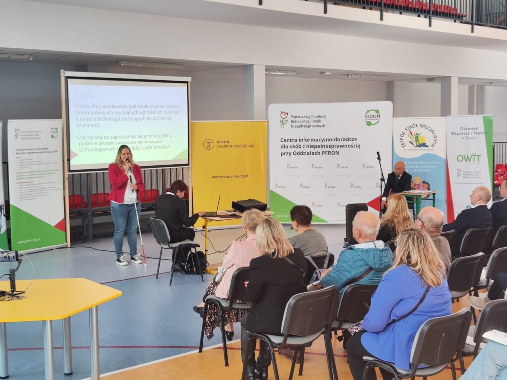 Na pierwszym planie trzy rzędy krzeseł z siedzącymi na nich osobami. W głębi ekran z prezentacją i rollupy. Przed nimi stoi pracownica OWiT w Opolu i mówi do mikrofonu.