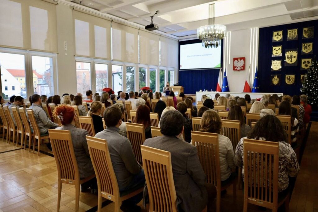 Na pierwszym planie około 80 osób uczestniczących w spotkaniu siedzi na krzesłach ustawionych w rzędy. Przed nimi stoi kobieta z mikrofonem w ręku. Za nią wisi ekran, obok godło Rzeczpospolitej oraz herb województwa opolskiego i herby powiatów województwa opolskiego.