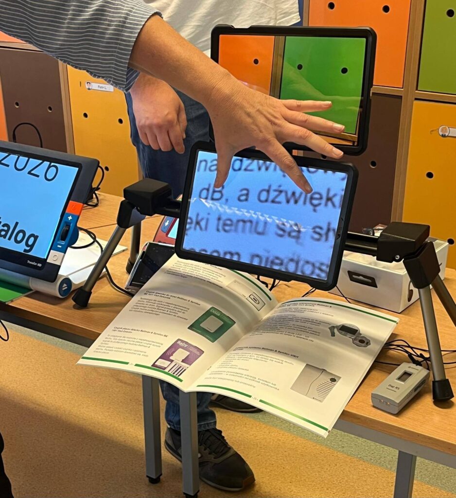 Dwa ekrany powiększalnika Exigo. Na pierwszym powiększony tekst z książki położonej pod ekranem. Na drugim ekranie powiększone szafki stojące w oddali.