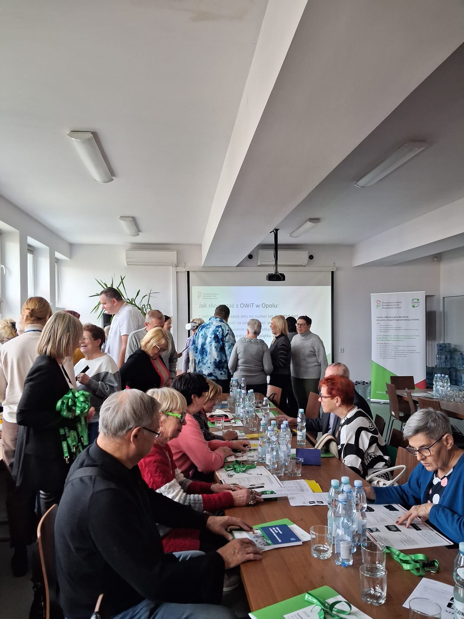 Na środku długi stół, po obu stronach siedzi 7 osób w różnym wieku. Na stole leżą katalogi technologii asystujących, smycze promocyjne PFRON, butelki z wodą i szklanki. Po lewej stronie stołu i za nim grupki około 15 osób w różnym wieku. Osoby rozmawiają ze sobą.
