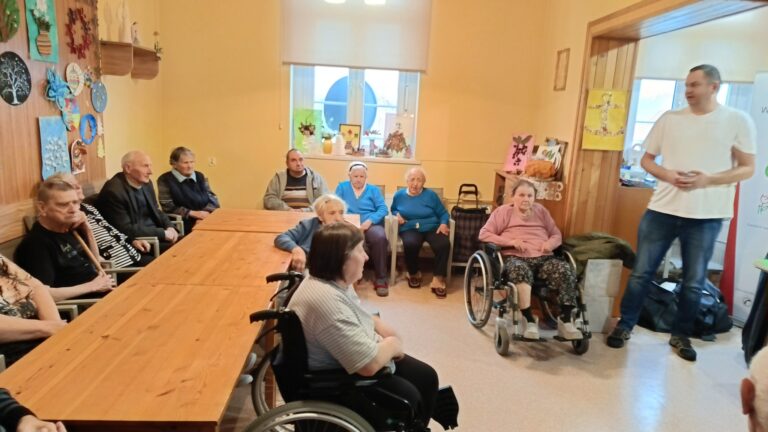 Po lewej stronie stoi długi stół. Po obu stronach stołu siedzi 11 osób. Dwie osoby siedzą na wózkach, pozostałe osoby siedzą na krzesłach. Po prawej stronie stoi pracownik Ośrodka Wsparcia i Testów w Opolu.