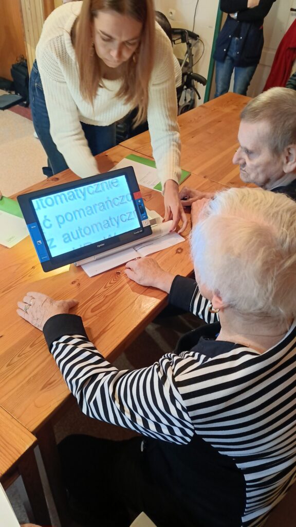 Przy stole siedzą kobieta i mężczyzna, oboje w wieku senioralnym. Przed nimi na stole stoi lupa elektroniczna Traveller. Za stołem stoi pracownica Ośrodka Wsparcia i Testów w Opolu. Pokazuje obsługę lupy.