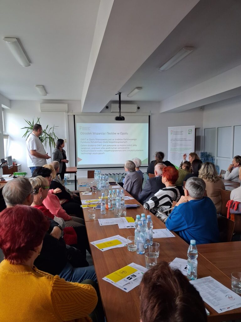 Na środku długi stół, po obu stronach siedzi 12 osób w różnym wieku. Na stole leżą ulotki PFRON dotyczące dostępności, butelki z wodą i szklanki. Po prawej stronie przy drugim stole siedzi 7 osób w różnym wieku. W głębi widoczny ekran z napisem „Ośrodek Wsparcia i Testów w Opolu”. Po lewej stronie ekranu stoi dwójka prelegentów: mężczyzna i kobieta.