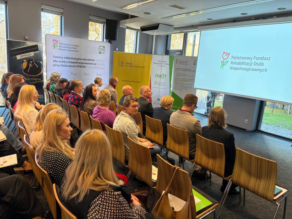 Sala konferencyjna. Na krzesłach ustawionych w rzędach siedzą uczestnicy spotkania. Przed nimi na ekranie wyświetlony napis Państwowy Fundusz Rehabilitacji Osób Niepełnosprawnych.