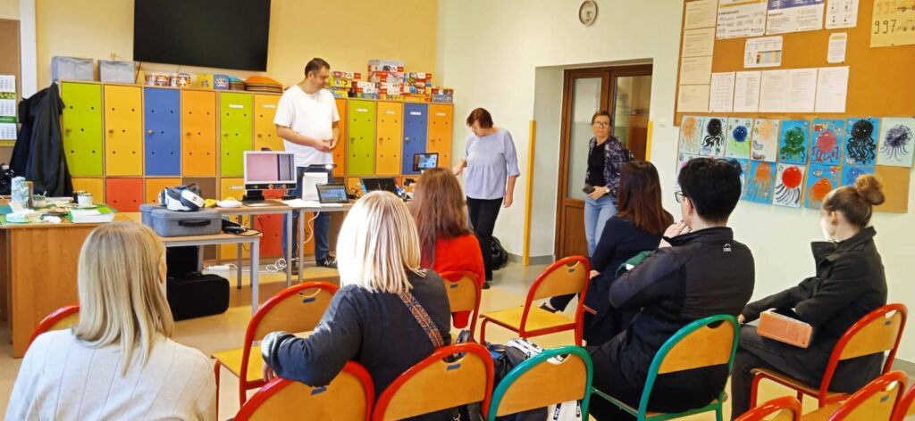 Sala świetlicy szkolnej. Grupa osób siedzi na krzesłach ustawionych w rzędach. Przed nimi na stołąch stoją technologie asystujące. Dwie osoby pracujące w OWiT prezentują technologie