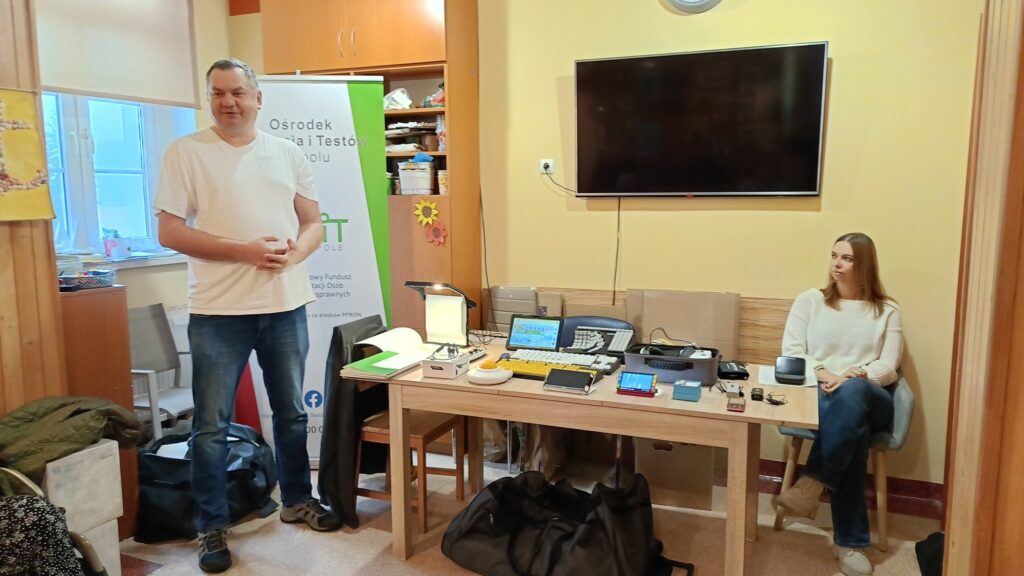 Na środku stoi stół. Na stole wystawione są technologie asystujące: lupy elektroniczne, klawiatury specjalistyczne, telefon udźwiękowiony, system FM, słuchawki kostne, duża mysz, tablet z Mówikiem, gogle relaksacyjne, urządzenie lektorskie. Po prawej stronie na krześle siedzi pracownica OWiT w Opolu. Po lewej stronie stoi pracownik OWiT Opolu.