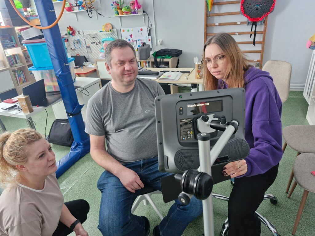 Kobieta i mężczyzna patrzą w monitor eytrackera, urządzenia sterowanego za pomocą wzroku. Po lewej stronie kuca kobieta, patrzy w monitor.