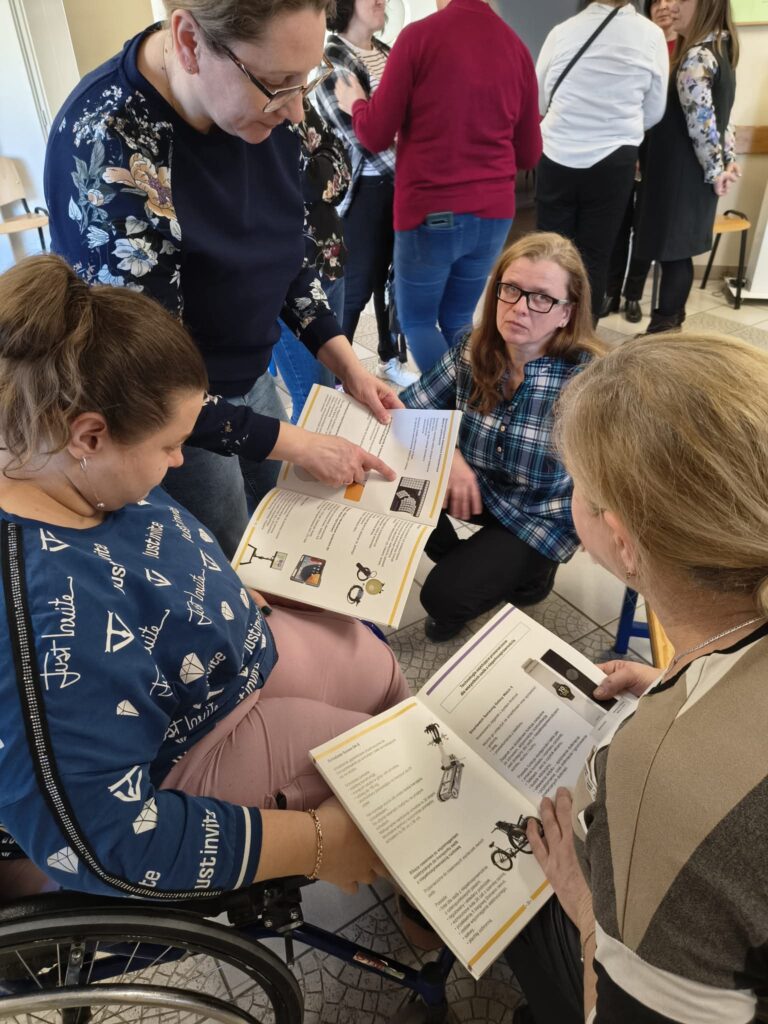 Cztery panie w różnym wieku w kręgu. Jedna stoi, jedna siedzi na wózku, jedna siedzi na krześle, jedna kuca. Dwie panie trzymają otwarte katalogi technologii asystujących.