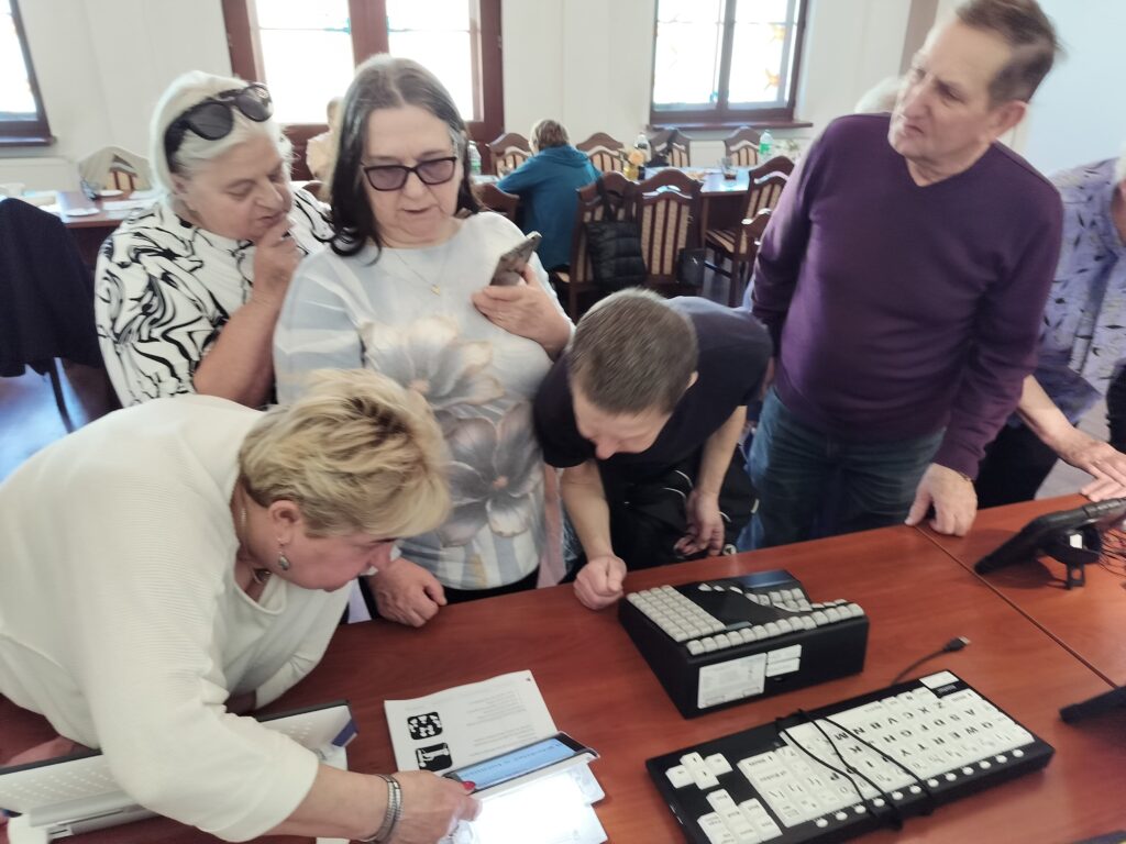 Trzy kobiety i dwaj mężczyźni stoją przy stole, na którym leży kartka z tekstem i obrazkami. Jedna z Pań trzyma w ręku lupę elektroniczną nad kartką. Pozostałe osoby przyglądają się kartce i lupie.