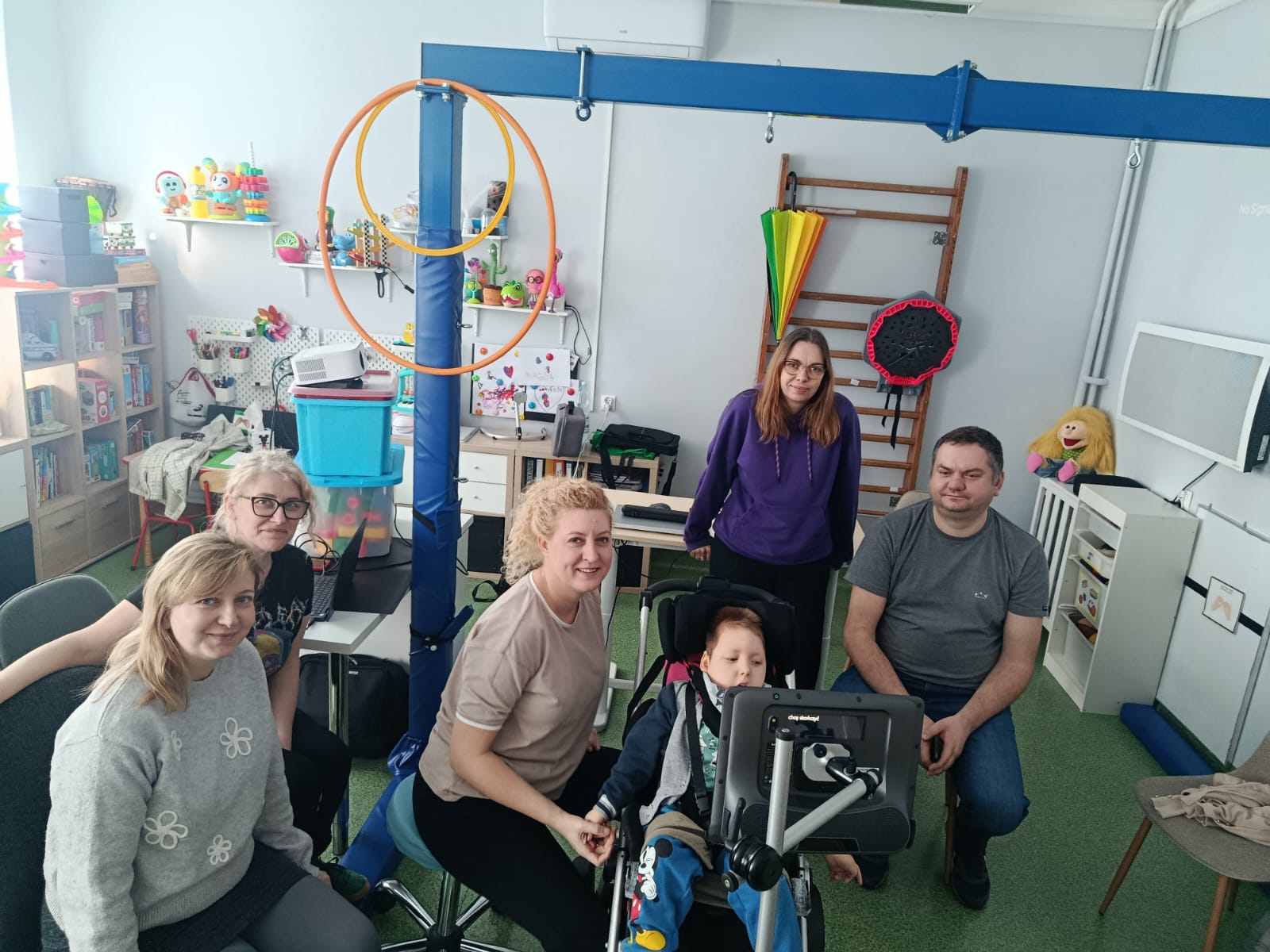 4 kobiety i mężczyzna oraz dziecko na wózku siedzą przez monitorem eytrackera, urządzenia sterowanego za pomocą wzroku.