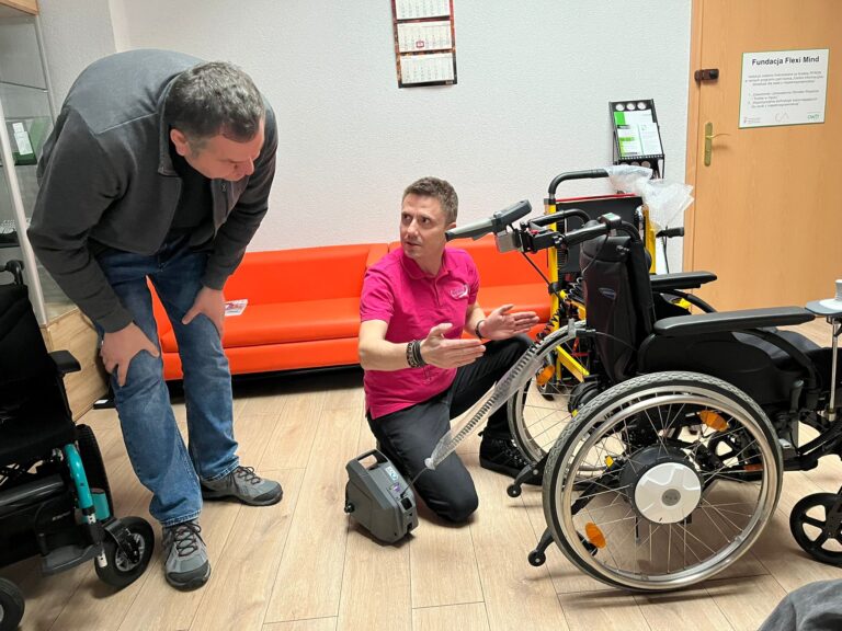 Po prawej stronie wózek z zamontowanym napędem e-fix. Obok wózka klęczy mężczyzna i rękami pokazuje jakąś odległość albo rozstaw. Po lewej stronie stoi nachylony pracownik OWiT i przygląda się wózkowi.