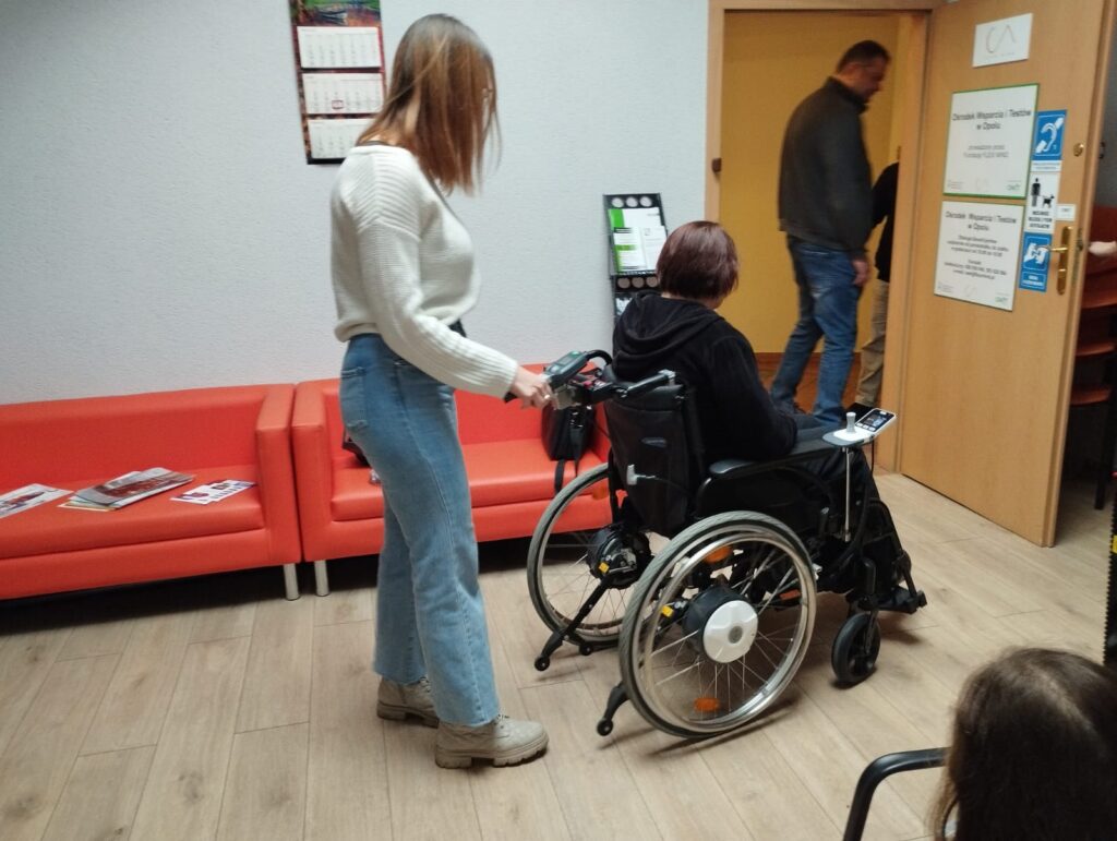 Na środku wózek z zamontowanym napędem e-fix. Na wózku siedzi kobieta. Po lewej stronie wózka stoi kobieta i uruchamia napęd z pozycji osoby prowadzącej wózek.