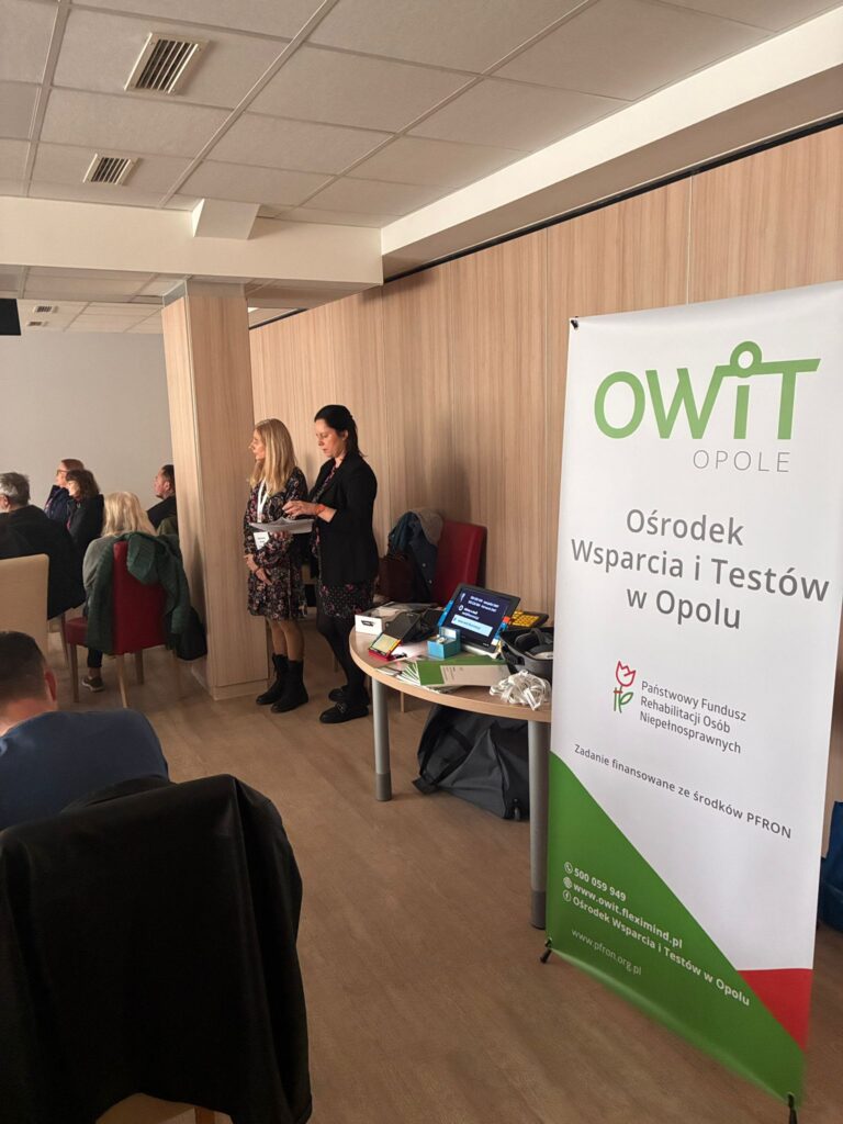 Po prawej stronie stoi banner OWiT w Opolu. Za nim stół z ułożonymi technologiami asystującymi, w tym lupa elektroniczna, powiększalnik, klawiatura z kontrastowymi klawiszami. Obok stoją dwie kobiety.