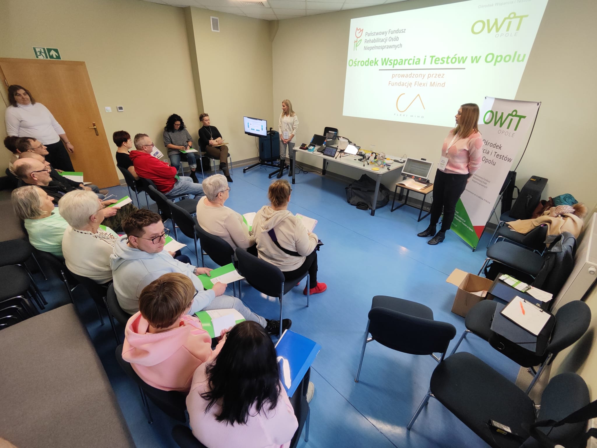 Na pierwszym planie widocznych jest od tyłu 16 osób siedzących na krzesłach. Krzesła ułożone są w dwa rzędy. Na drugim planie na ścianie duży obraz z rzutnika z napisem Ośrodek Wsparcia i Testów w Opolu prowadzony przez Fundację Flexi Mind. Obok napisu jest logo PFRON, OWIT Opole i Fundacji Flexi Mind. Na drugim planie na środku sali środku stoją dwie pracownice OWiT Agnieszka i Kasia.