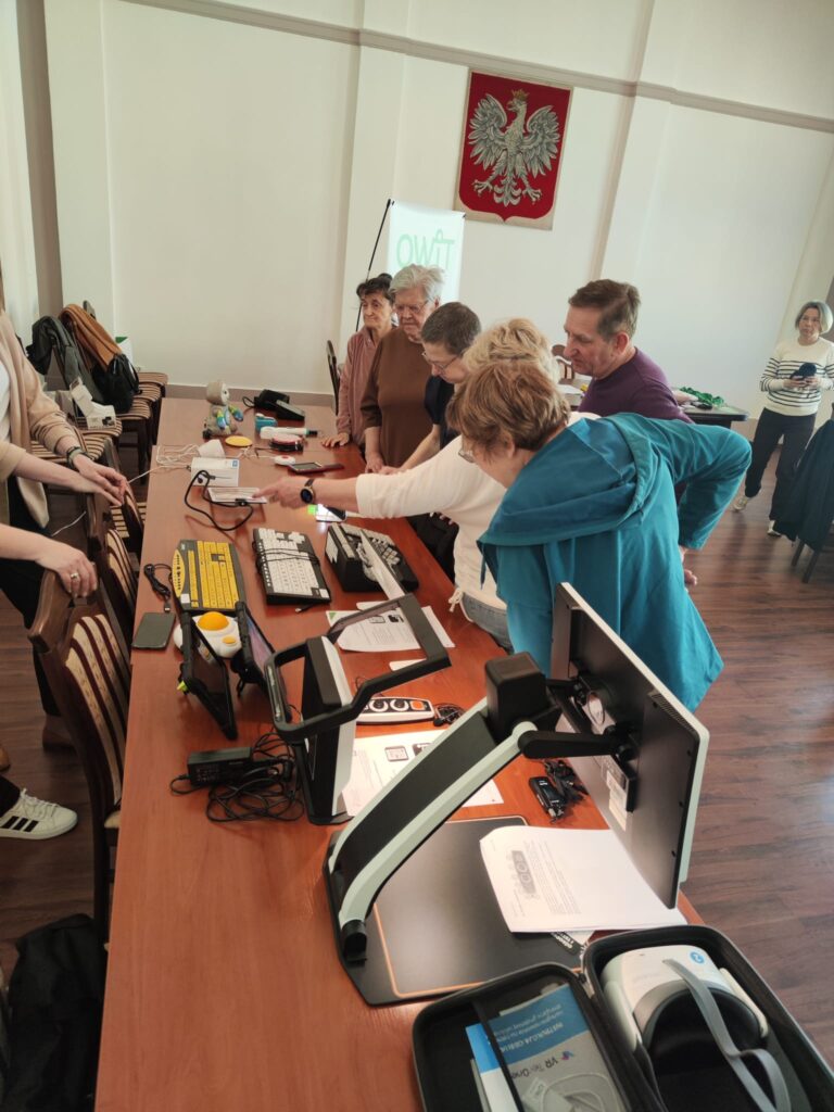 Na długim stole rozłożone są technologie asystujące między innymi: powiększalnik, urządzenie lektorskie, klawiatury z klawiszami w kontrastowym kolorze, powiększona mysz komputerowa, mówiący ciśnieniomierz, udźwiękowiony zegarem na rękę. Po prawej stronie stołu stoi sześć osób w różnym wieku i różnej płci, oglądają zgromadzone technologie.