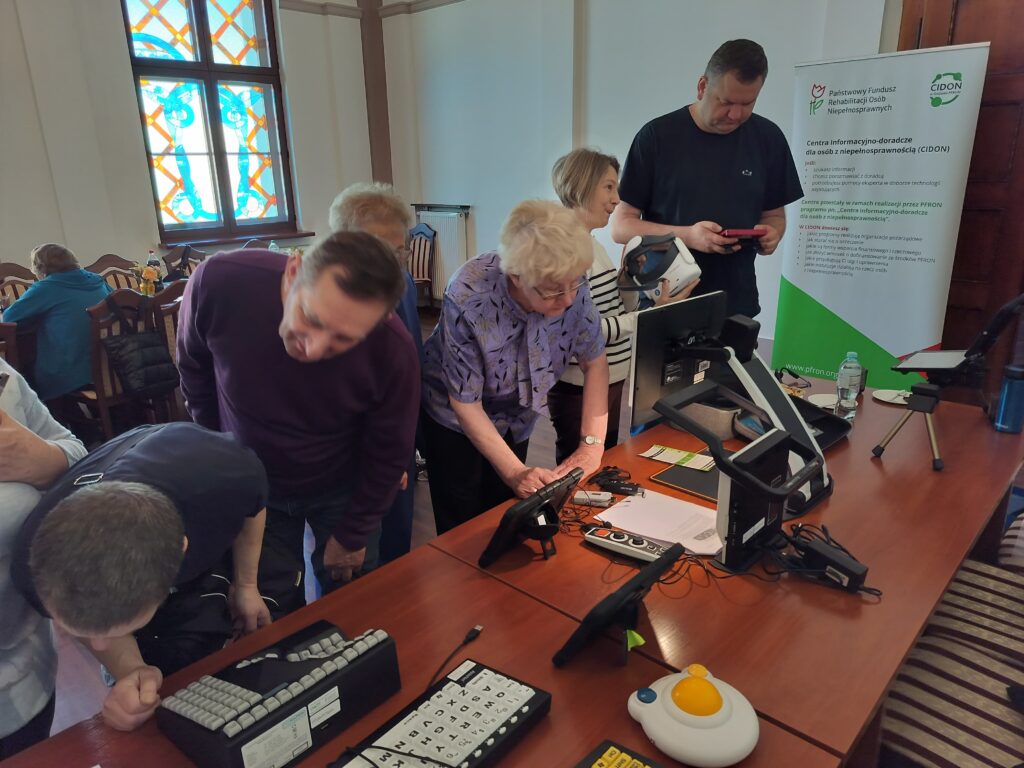 Na długim stole rozłożone są technologie asystujące między innymi: powiększalnik, urządzenie lektorskie, klawiatury z klawiszami w kontrastowym kolorze, powiększona mysz komputerowa, mówiący ciśnieniomierz, udźwiękowiony zegarem na rękę. Po lewej stronie stołu stoi siedem osób w różnym wieku i różnej płci, oglądają zgromadzone technologie.