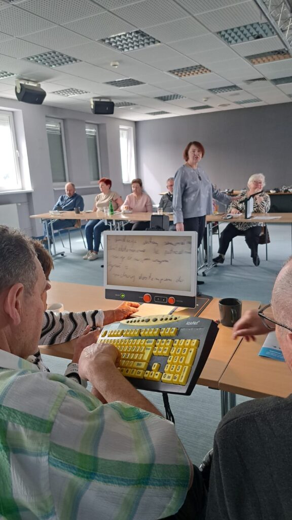 Przy stole tyłem do aparatu siedzi mężczyzna. Trzyma klawiaturę z kontrastowymi klawiszami. Przed nim stoi powiększalnik. W tle na środku sali stoi osoba prowadząca spotkanie. Za nią przy kolejnym stole siedzi dwóch mężczyzna i dwie kobiety. Na stole stoi urządzenie lektorskie.